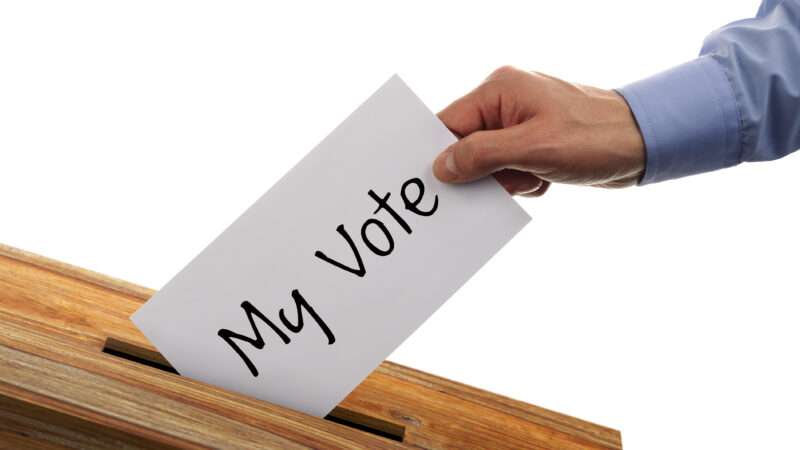 A hand places a white piece of paper, which says "my vote," into a box.