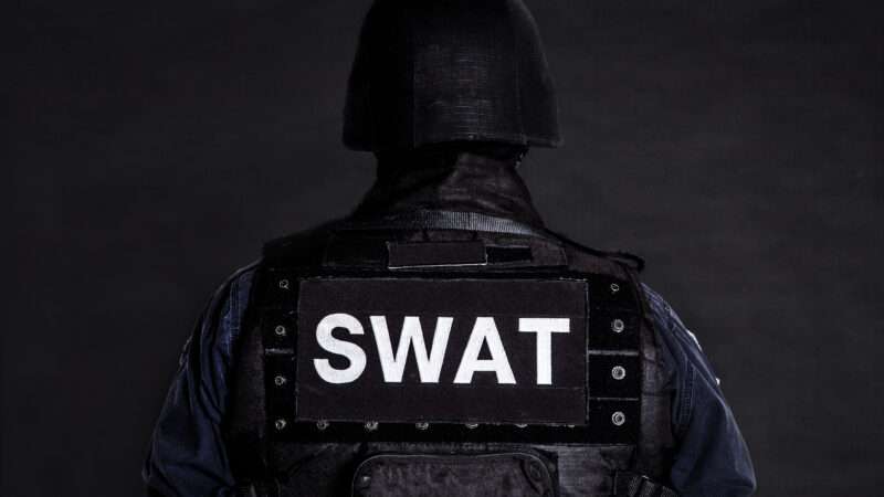 A SWAT officer faces away from the camera against a dark gray background.
