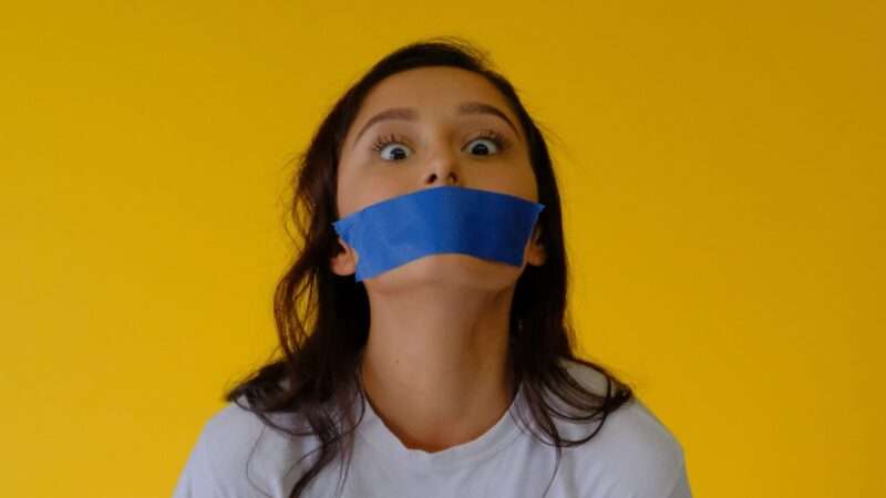 A young woman with blue tape over her mouth against a yellow background