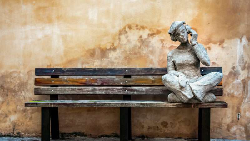 A stone statue of a young person listening to music, placed on a wooden bench against a stone wall. | Cinar Yilancioglu | Dreamstime.com