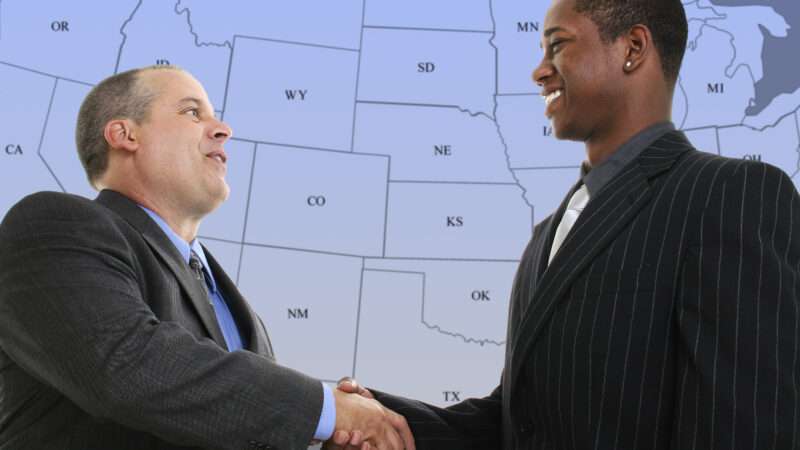 Two businessmen shaking hands in front of a blue and grey map of the United States.