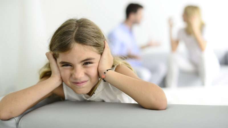 Exasperated, a little girl clamps her hands over her ears as her parents argue in the background.