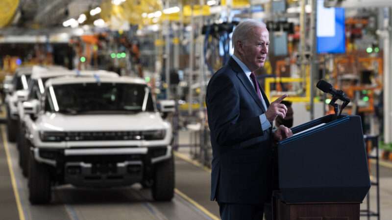 President Joe Biden speaks in front of a lectern inside an automobile factory, with a line of Hummer EVs in the background.