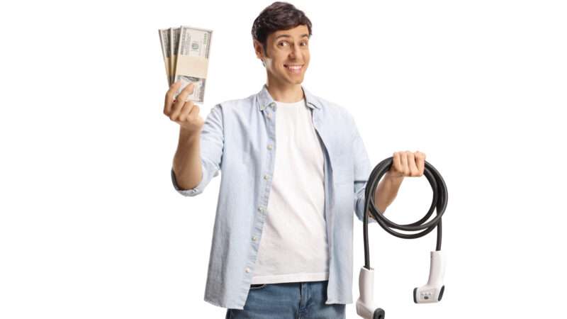 A man holds an electric vehicle charging cable in one hand and a fanned-out stack of cash in the other. | Ljupco | Dreamstime.com