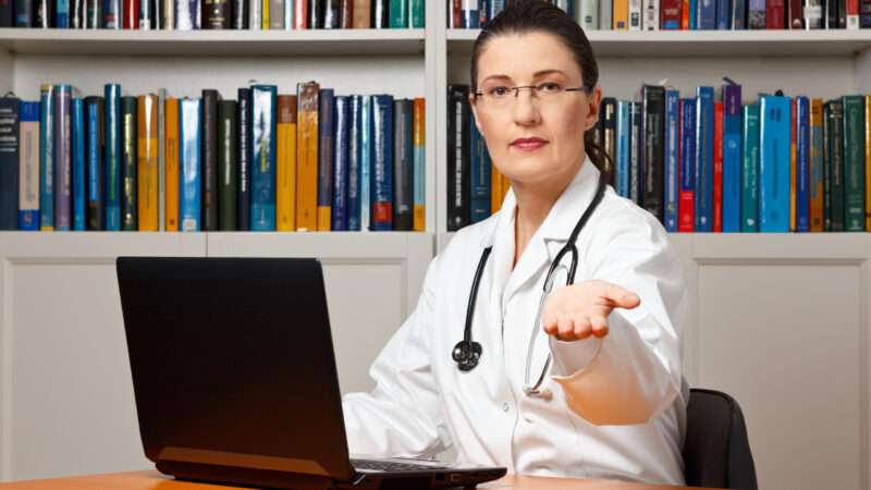 Female doctor sitting in front of a bookcase and behind a laptop with her hand sticking out, as if to ask for money. | Photo 63906299 © Agenturfotografin | Dreamstime.com