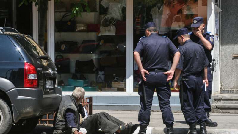 Police officers surround a homeless man leaned against a car. | Gabriel Petrescu | Dreamstime.com