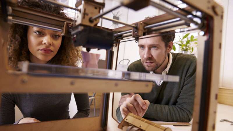 Two people peer into a 3D printer. | Monkey Business Images | Dreamstime.com