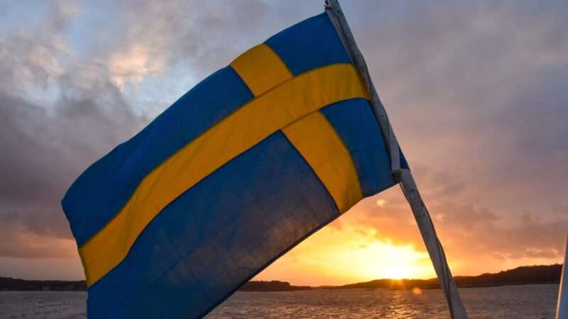 A Swedish flag waving in the wind in front of a sunset. | Photo by <a href="https://unsplash.com/@simonclare1?utm_content=creditCopyText&utm_medium=referral&utm_source=unsplash">Simon Clare</a> on <a href="https://unsplash.com/photos/a-blue-and-yellow-flag-on-a-boat-in-the-water-zx8PwNg2wuI?utm_content=creditCopyText&utm_medium=referral&utm_source=unsplash">Unsplash</a>
