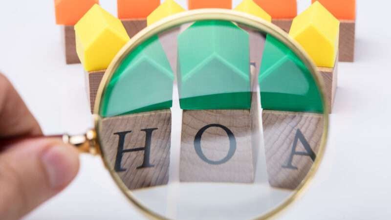 A magnifying glass in front of wooden letters spelling HOA and a row of multicolored houses behind it against a white background | Andrey Popov/Dreamstime.com