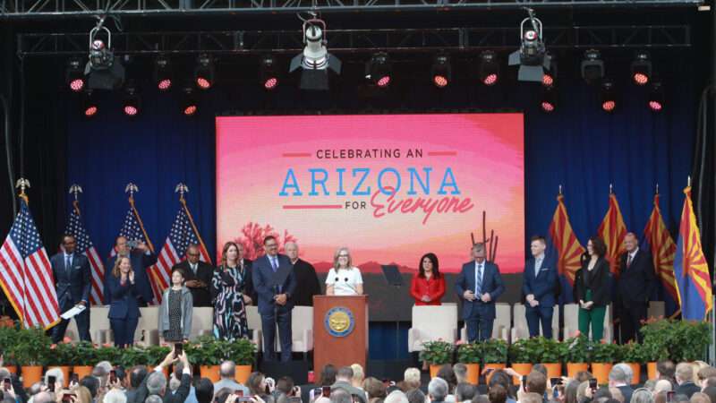 Elected officials in Arizona stand on a stage in front of a screen that reads, 