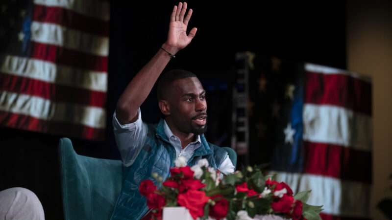 The activist DeRay Mckesson, in his trademark blue Patagonia vest, waves from a stage in front of American flags. | Bob Daemmrich/ZUMA Press/Newscom