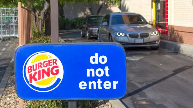 The end of a Burger King drive-thru, showing cars in line and a sign with the franchise logo that says 