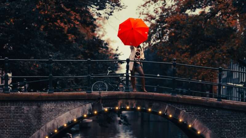 Woman standing on a small bridge, holding a red umbrella over her face | Photo by <a href="https://unsplash.com/@driftinghorizon?utm_content=creditCopyText&utm_medium=referral&utm_source=unsplash">Peyman Shojaei</a> on <a href="https://unsplash.com/photos/a-person-holding-an-umbrella-over-a-bridge-635eTFNeKjU?utm_content=creditCopyText&utm_medium=referral&utm_source=unsplash">Unsplash</a>       