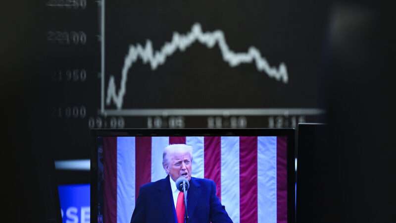 Donald Trump speaks in front of an American flag, above him is seen a black and white image of a stock line trending downward | Arne Dedert/dpa/picture-alliance/Newscom