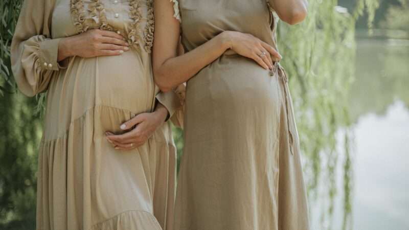 Two pregnant women stand next to one another outside. Their heads are cropped out of the image, they are both wearing tan dresses, and they each have hands resting on their stomachs | Photo by <a href="https://unsplash.com/@yuli_superson?utm_content=creditCopyText&utm_medium=referral&utm_source=unsplash">Juli Kosolapova</a> on <a href="https://unsplash.com/photos/woman-in-white-floral-dress-lM2dFjHccJs?utm_content=creditCopyText&utm_medium=referral&utm_source=unsplash">Unsplash</a>       
