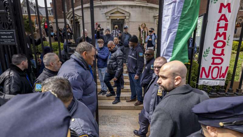 People in law enforcement standing in front of a fence with banners hanging from it, with people in the background, on the other side of the fence, holding up phone cameras. |  Michael Nigro/Sipa USA/Newscom