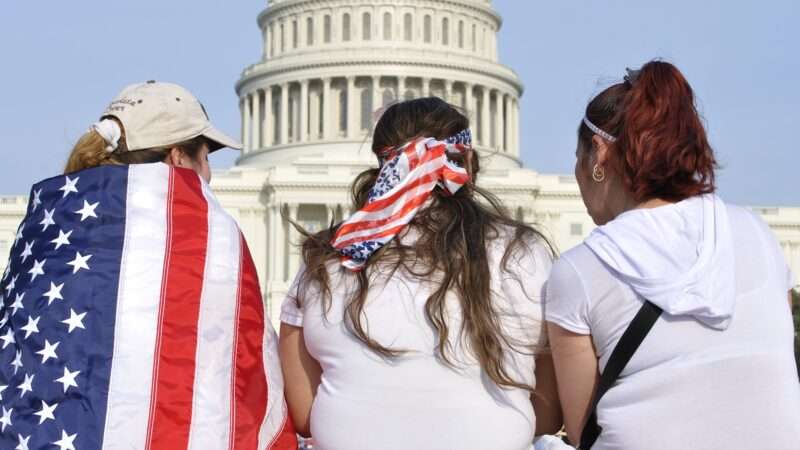 Three women sit with their backs to the camera, facing the U.S. Capitol. One woman has an American flag around her shoulders, while another has her hair in an American flag print bandana. | Czuber | Dreamstime.com