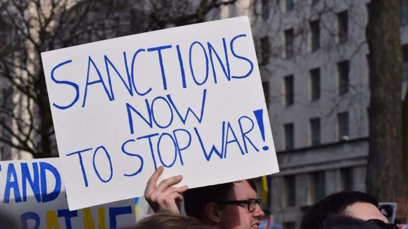 A protester holds a pro-sanctions sign that reads SANCTIONS NOW TO STOP WAR! in blue lettering on a white background | ID 242338731 ©  Amani A | Dreamstime.com