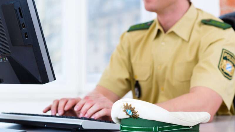 A policeman sits, with his hat on the desk, happily typing on a computer. | Arne9001 | Dreamstime.com