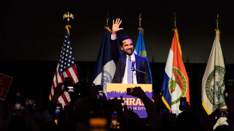 Zohran Mamdani speaks to a crowd behind a podium with his campaign sign attached and with flags in the background | Lev Radin/Sipa USA/Newscom