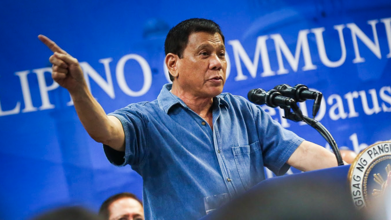 Rodrigue Duterte giving a speech in front of a lectern with two microphones, wearing a blue denim shirt, with his right hand pointing to the side. | Wikimedia Commons