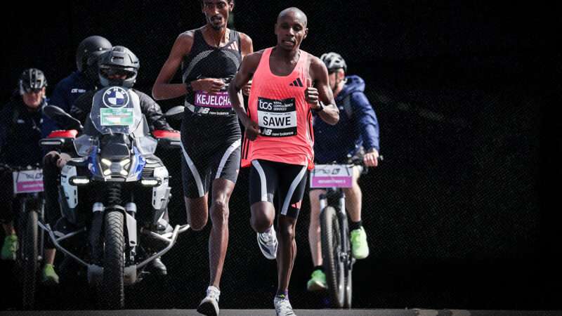 In front of a black background, Sabastian Sawe and Yomif Kejelcha run toward the camera as two bicycles and two motorcycles trail behind them. | Credit: Henning von Jagow / Action Plus/Newscom