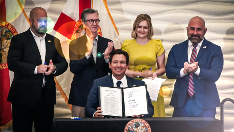 Gov. DeSantis holds a legal document and smiles from behind a desk with people standing behind him | Governor Ron DeSantis/Youtube