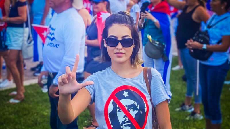 A woman wearing sunglasses and a t-shirt with the red "ban" symbol over Che Guevara's face is holding up her hand in the shape of an L. | Germania Rodríguez Poleo