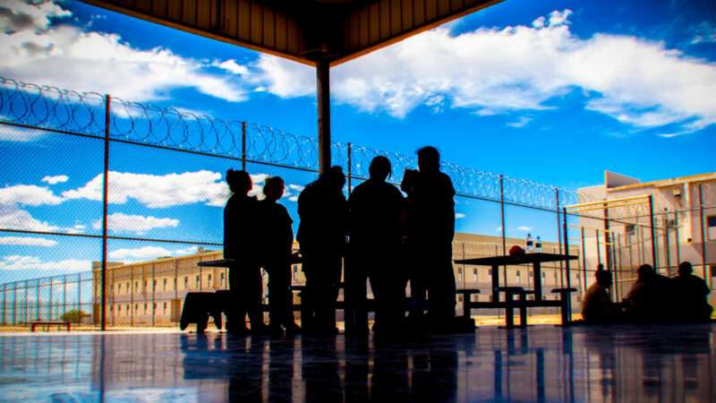 A group of people huddled in the shade beneath a roof, standing next to a barbed wire fence. | Photo: U.S. Immigration and Customs Enforcement