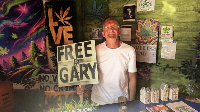 A man stands behind a counter in a plain white t-shirt and glasses holding a sign that says "FREE GARY." The walls and counter are filled with marijuana imagery. | Mattha Busby
