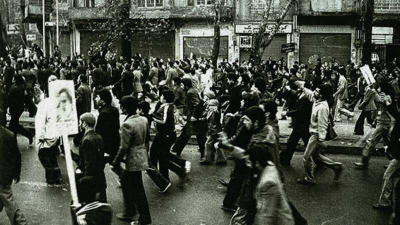 A black-and-white photo of people marching in a street, all toward the left side of the photo frame. A few of them hold signs on posts. | Wikimedia Commons