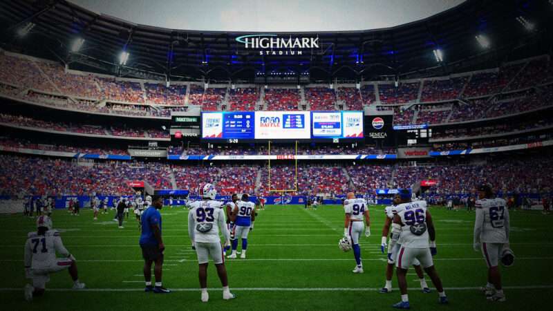 A group of players in football jerseys stands on the field, spread out, all facing a far-off end zone. The stands rise above them with speckles of red, white, and blue, with a video scoreboard and a logo that says
