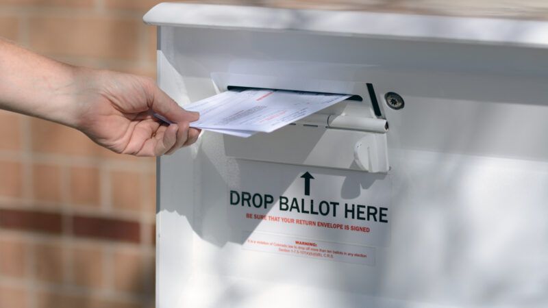 A hand places a ballot into a ballot drop-box.