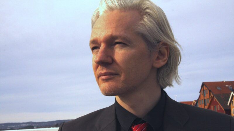 Julian Assange in a suit looking to the left of the camera with a gray sky behind him.