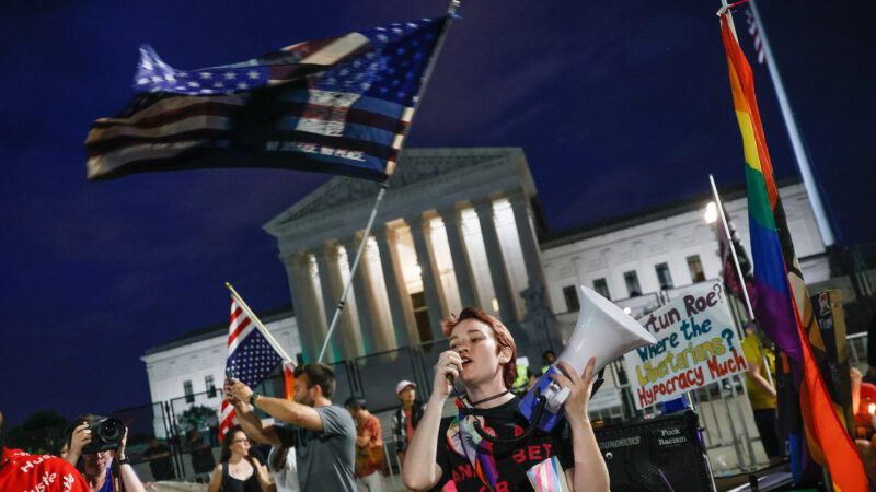 Protestor speaking into a megaphone outside the Supreme Court at night while another protestor waves an American flag.