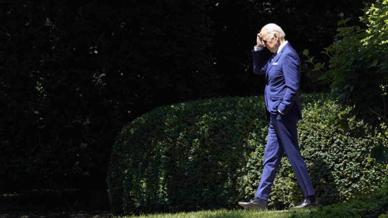 President Joe Biden walking outside past shady bushes and trees.