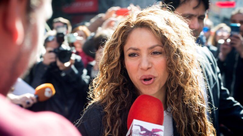 A blonde-haired woman with curly hair speaks to a reporter holding a red microphone