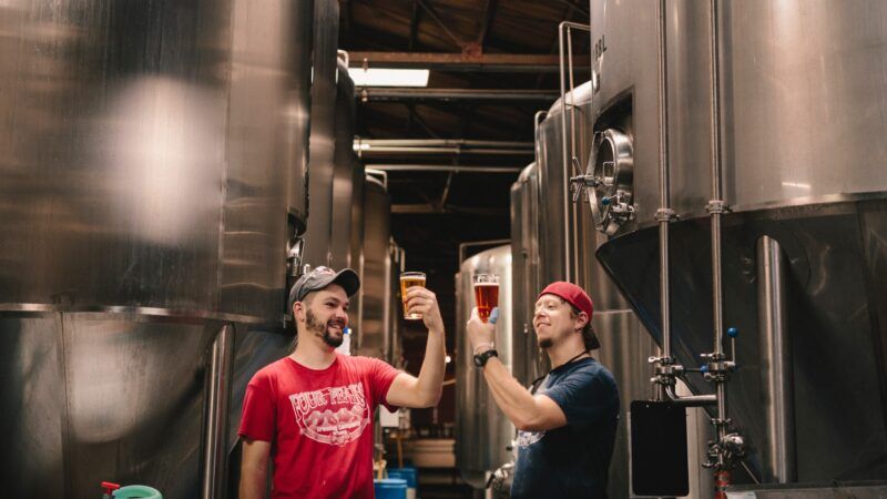 Two men holding full beer glasses up amid tall stainless steel tanks in a brewery. | Photo by <a href="https://unsplash.com/@elevatebeer?utm_source=unsplash&utm_medium=referral&utm_content=creditCopyText">Elevate</a> on <a href="https://unsplash.com/s/photos/brewery?utm_source=unsplash&utm_medium=referral&utm_content=creditCopyText">Unsplash</a>