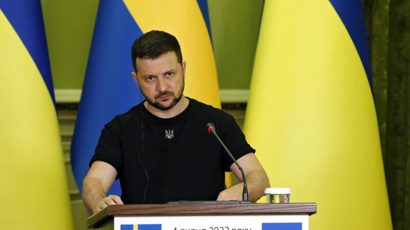 Ukrainian President Volodymyr Zelenskyy in a t-shirt at a podium with three Ukraine flags behind him.