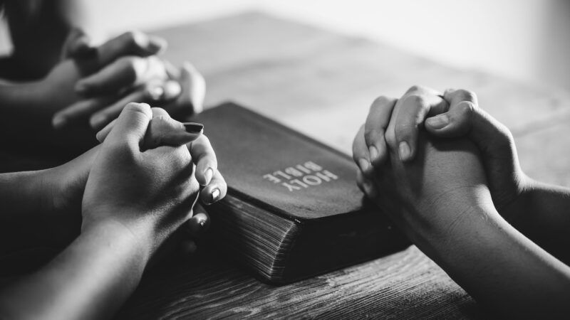 clasped hands rest on a table around a copy of the Bible