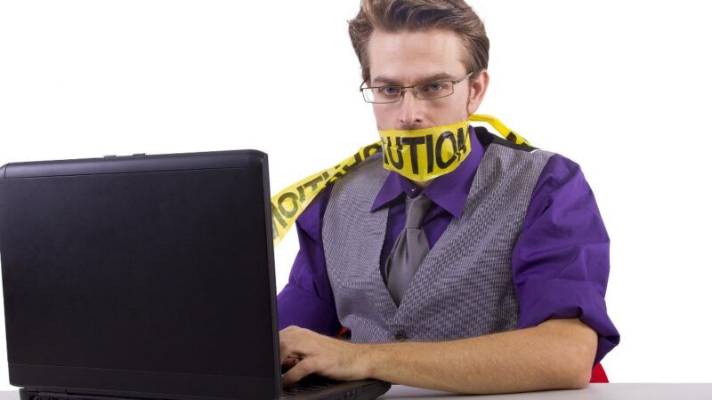 Man with yellow tape wrapped around his mouth while on a laptop | Innovatedcaptures / Dreamstime.com