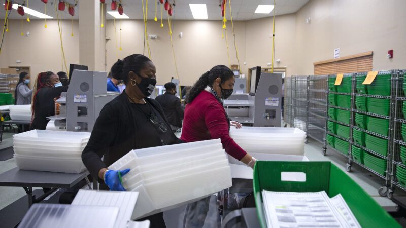 Two elections workers wearing face masks process bins full of ballots in an elections office