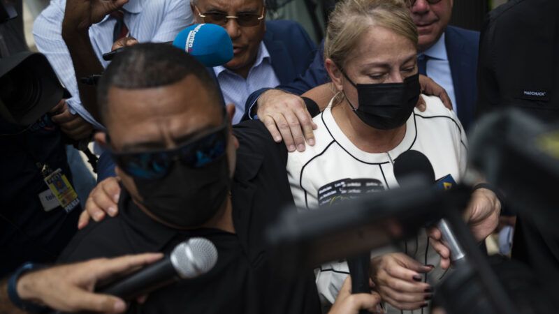 Former Governor of Puerto Rico Wanda Vazquez, a woman wearing a white and black blouse with a black face mask on, flanked by attorneys and reporters after being arrested.