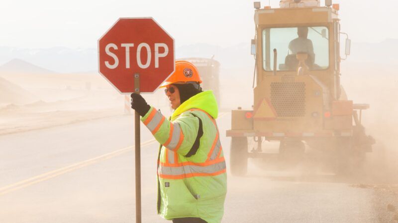 A construction worker holds a STOP sign with dust and road work in the background
