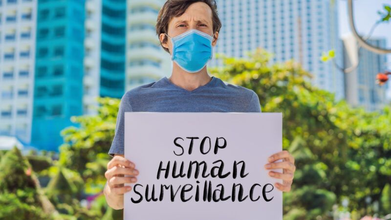 a masked man standing in front of skyscrapers holding a sign reading "stop human surveillance"
