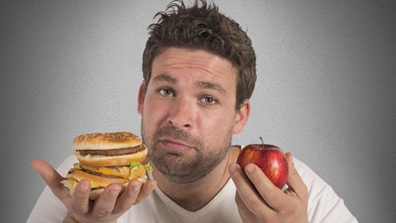 A disheveled man holds a hamburger and an apple.