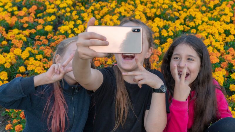 Three teen girls take a smiling self on an iPhone in front of a field of sunflowers.