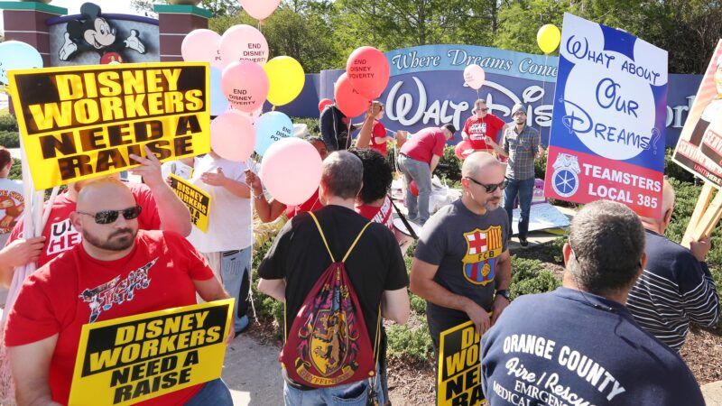 A crowd protests Disney with placards saying "DISNEY WORKERS NEED A RAISE"