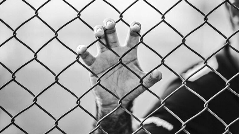 man behind prison fence | Photo by <a href=https://reason.com/2023/02/28/prison-deaths-spiked-by-almost-50-percent-during-early-months-of-covid-19-pandemic/