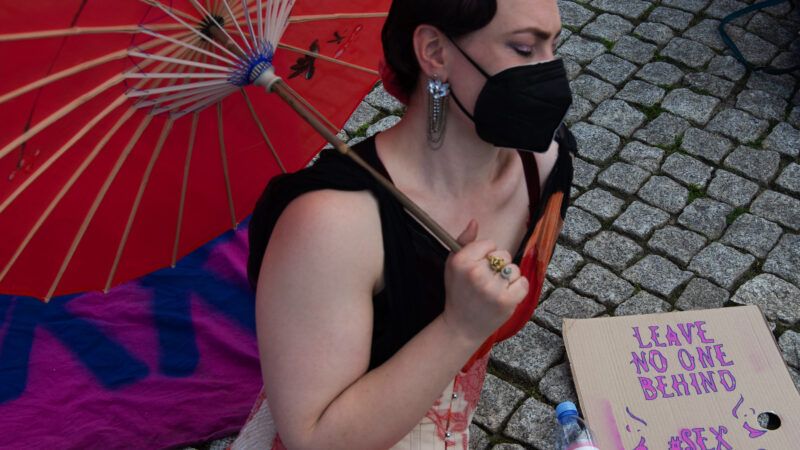 protester with red umbrella and a sign saying "leave no one behind | sex work is work"